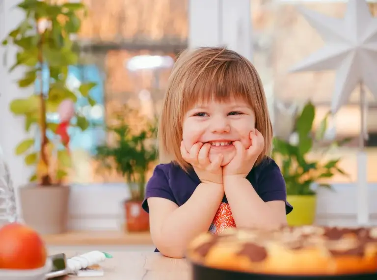 A young girl with her hands under her chin smiling, blurred background for focus on girl.