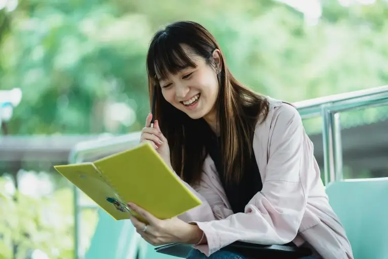 Teen reading a book sitting on a picnic table and smiling.